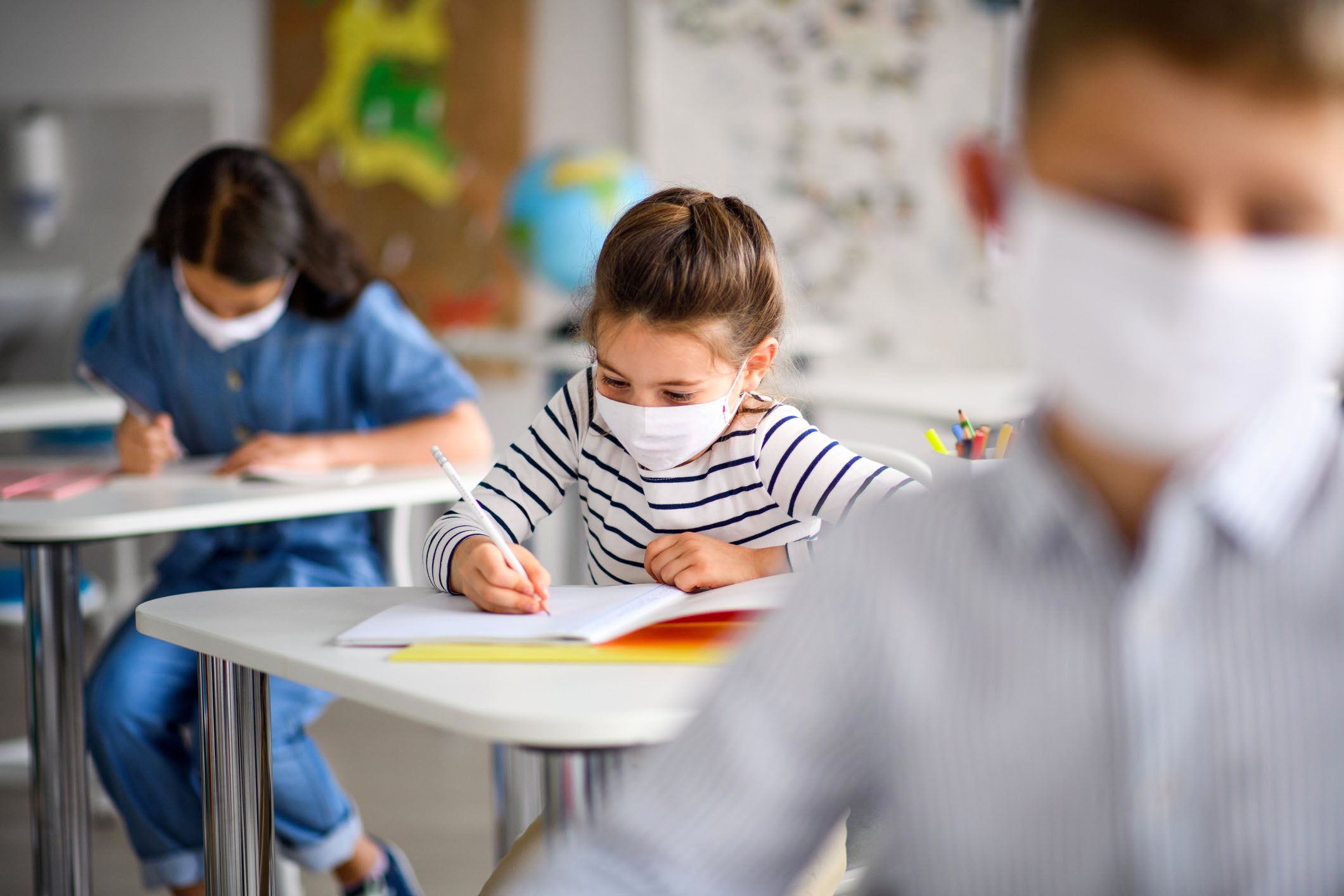 Young children wearing face masks sitting at desks writing in a classroom.